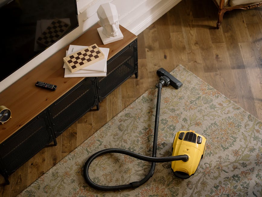 A vacuum cleaner is being used on an ornate, patterned carpet in a residential living room, with a person standing nearby, wearing sneakers and dark pants. The vacuum head is black with a control swit
