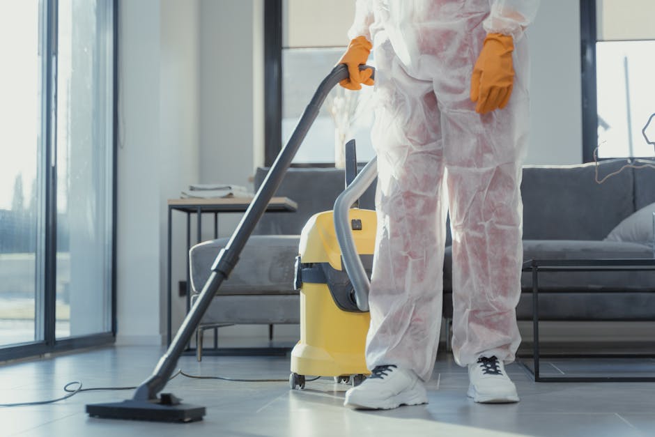 Close-up of a professional cleaner dressed in a white protective suit and blue gloves, holding a spray bottle with a blue liquid and a cleaning cloth. The background shows a dining area with a wooden